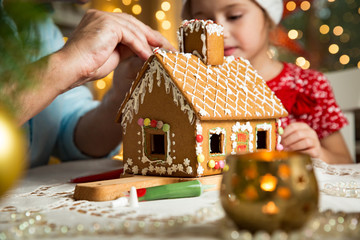 Father and adorable daughter in red hat building gingerbread house together. Beautiful decorated...
