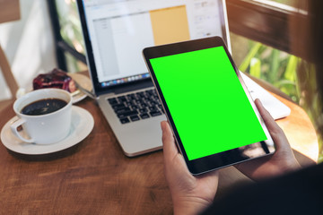 Mockup image of hands holding black tablet pc with blank green screen , laptop , coffee cup and cake on wooden table in cafe