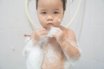 Little asian girl taking shower in bathroom, playing bath foam bubbles. Selective focus