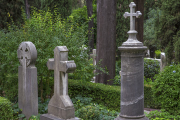 Dettaglio di tre croci poste sopra le rispettive tombe in un cimitero cattolico italiano. Le croci sono costruite in pietra in mezzo a tanti alberi.