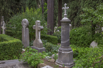 Dettaglio di tre croci poste sopra le rispettive tombe in un cimitero cattolico italiano. Le croci sono costruite in pietra in mezzo a tanti alberi.