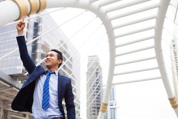 Businessman smiling and raising his fist with number 1 sign in the air, with office building background - business success, achievement, and win concepts