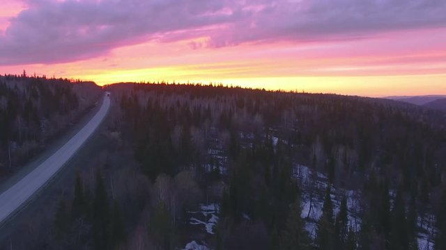 AERIAL: The camera flies over a thick forest with bloody red sunset and mountains on a background. Nearby there is a curved road with a lonely traveling truck. Early winter with some snow.
