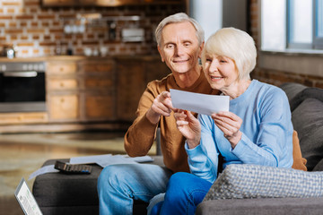 Happy couple. Positive nice aged couple sitting on the sofa and looking at the piece of paper while...