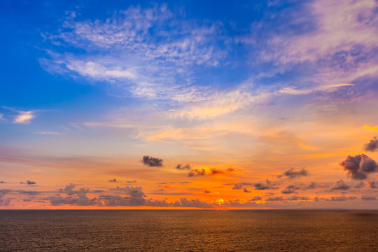 Bright colorful sun hides behind orange clouds, goes beyond the horizon of the sea, the blue yellow sky above the Indian Ocean. Beautiful sunset from Uluwatu Temple, Bali, Indonesia.