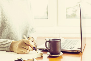 woman writing in a notepad sitting at her workplace by the window with a laptop and a cup of coffee