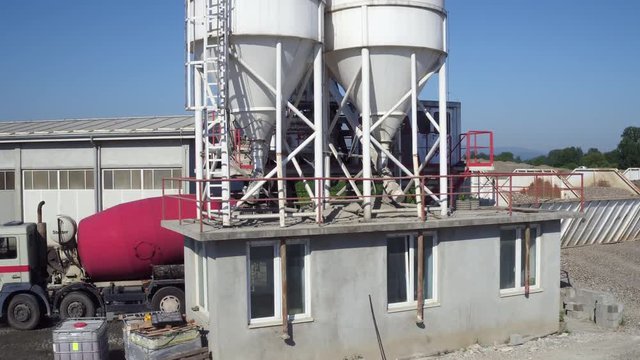 Mixer truck loading concrete from cistern, aerial view, camera motion over tank to countryside landscape with villages, mountain, forest and fields, bulldozer pushing sand on heap, building industry