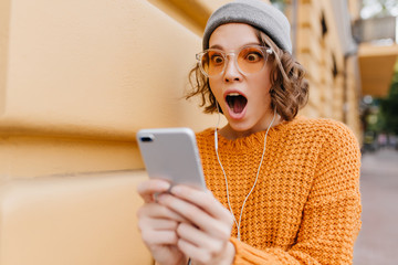 Shocked girl with brown eyes looking at phone screen with mouth open. Outdoor portrait of surprised...