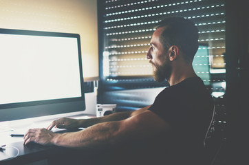 A bearded caucasian man is sitting behind the blank white monitor of the computer. Muscular handsome guy is looking at the blank display during working time. Empty space on the pc screen.