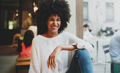 Close up portrait of a beautiful cheerful afro-american woman with black curly hair wearing stylish clothes looking at the camera while sitting in a modern city coffee shop on a weekend day.