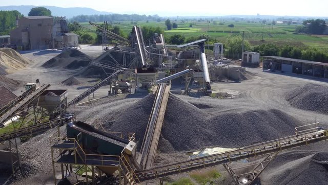 Processing plant, stone crusher machines in work process, conveyor belts transporting rocks and ground, tipper truck passing, gravel and sand production, aerial view, back light, sunny day