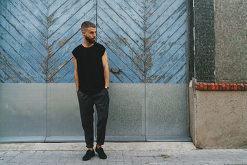 Full length portrait of a handsome bearded man dressed in fashionable clothes standing on a street against the retro light blue wooden gates. Handsome hipster guy posing in a blank black t-shirt.