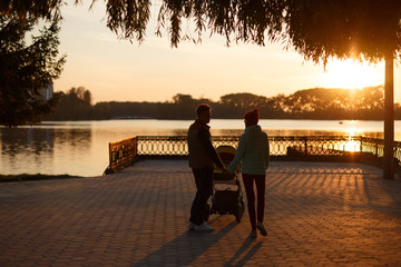 Back view of young loving couple silhouettes holding hands. Parents with baby pram in autumn park...