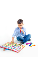 A cute cheerful boy in a blue checkered shirt and jeans with an alphabet and colored felt-tip pens sits on a white background