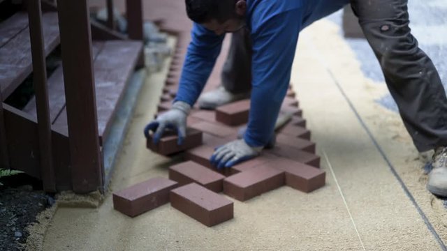 Man putting red brick pavers into place in a herringbone pattern on a bed of sand.
