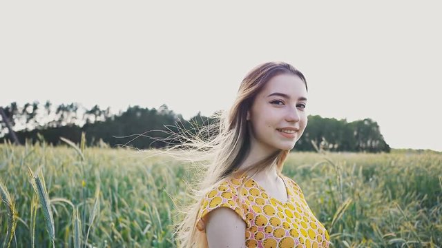 A beautiful romantic girl walking alone through a field of green wheat and touching wheat ears. She corrects long hair and enjoys nature on a summer sunny day.