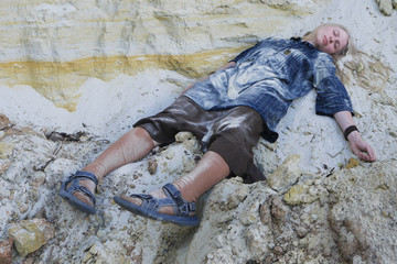 A girl lies on the sand near the sandstone wall with eyes closed and head on side. The young woman is dressed in blue shirt, brown skirt, sandals. She acts like been killed, fallen to death or asleep