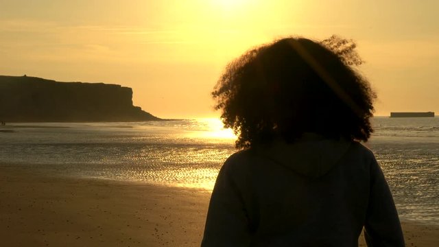 4K video clip of sad thoughtful mixed race African American girl teenager female young woman with curly hair standing on a beach looking at sunset or sunrise