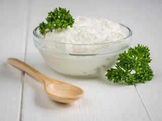 Curly leaves of parsley and a bowl of curd cream on a white table.