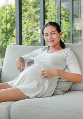 Pregnant woman is siting on sofa and is putting headphones on her belly.