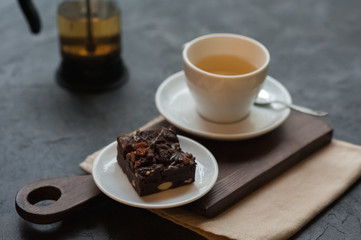 Morning photo, breakfast on a dark concrete background. White cup on white saucer with tea. A piece of chocolate brownie on a plate on a wooden board. Tea fountain- press. Tablecloth in the background