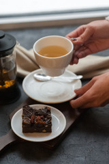 Morning photo, breakfast on a dark concrete background. White cup on white saucer with tea. A piece of chocolate brownie on a plate. Tea fountain - press. A human hand holds a plate with a cake.