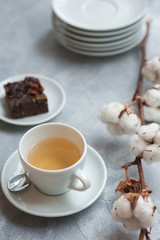 Morning photo, breakfast on a light concrete background. White cup on white saucer with tea. A piece of chocolate brownie on a plate. Sprig of dried cotton.