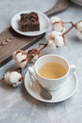 Morning photo, breakfast on a light concrete background. White cup on white saucer with tea. A piece of chocolate brownie on a plate. Sprig of dried cotton.