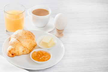 Continental breakfast with a croissant, boiled egg. Coffee or tea with milk, a glass of juice, buns, butter, jam. Soft backlight.  From above