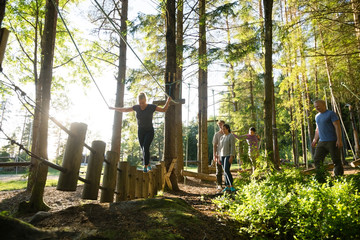 Friends Watching Woman Crossing Log Bridge In Forest