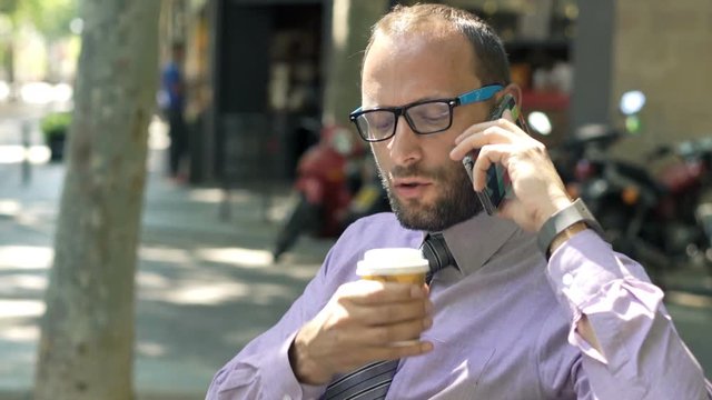 Young businessman with coffee talking on cellphone sitting on bench in city
