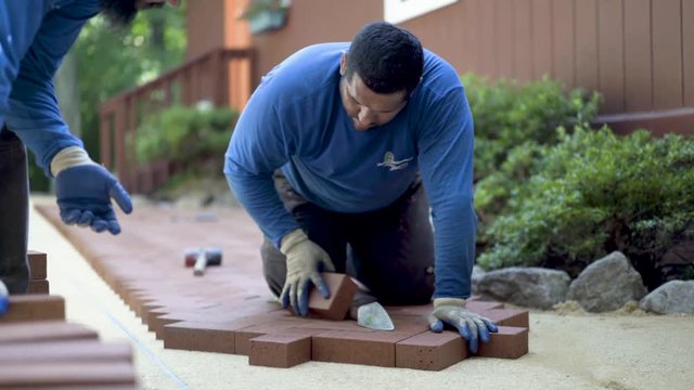 One man hands red bricks to another man who is putting the brick pavers into place in a herringbone pattern.