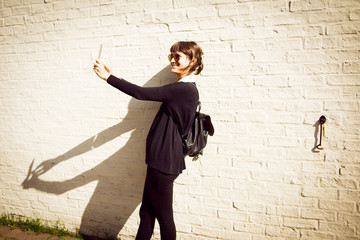 Young woman dressed in black with a small leather backpack wearing sunglasses smiling and taking a selfie in front of a white brick wall as background - 179953246