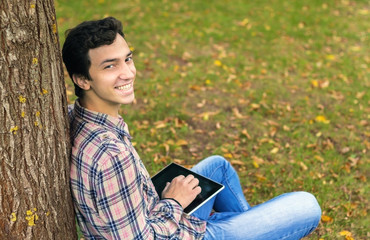 young men student sitting  in the park next to the tree with the tablet in his hands