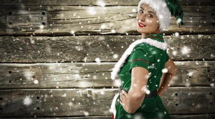 Piękna młoda kobieta w kostumie swiątecznego elfa.  Zielona sukienka i czapka. Na tle drewnianej sciany. Sypiący snieg .  - obrazy, fototapety, plakaty