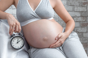 Pregnant woman holding a clock. Close up belly in underwear. Waiting for birth. Motherhood concept.