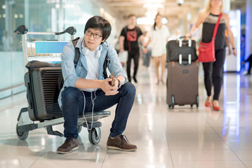 Young Asian man sitting on airport trolley with his suitcase luggage in the international airport terminal, waiting for travel abroad