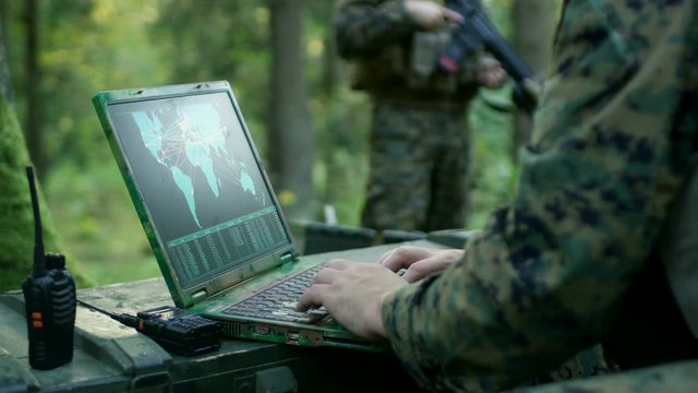 Military Operation in Action, Soldiers Using Military Grade Laptop Use Military Industrial Complex Hardware for Accomplishing International Mission. In the Background Camouflaged Tent on the Forest. 