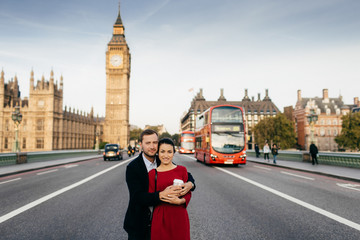 Outdoor shot of romantic couple embrace each other, stand on Westminster Bridge, have excursion, pose at camera against Big Ben and busy city scene with transport, enjoy being in London