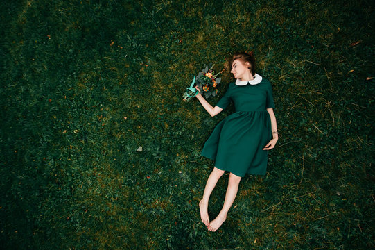 Young Woman Portrait From Above. Beautiful Irish Cute Girl With Red Hair Lying On Grass With Bouquet Of Flowers In Hand. Minimalism. Fairy Tale And Fable. Odd Strange Bizarre Female With Naked Legs.