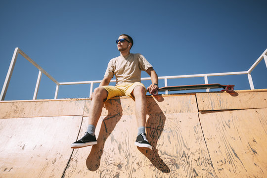 Young man with earbuds and longboard sitting on top of halfpipe in skatepark