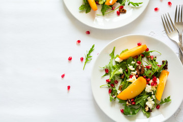 Fresh autumn salad with persimmon, arugula, cheese and pomegranate seeds on white table cloth. Space for copy