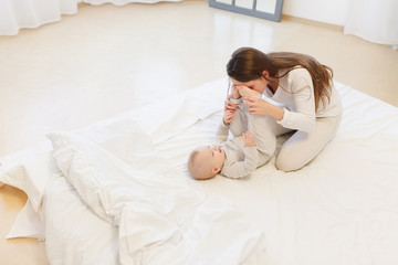 Happy cheerful young mom smiling playing with her little baby boy lying in bed at home. family portrait, white clothes, light house interior