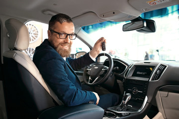 Formal wearing young man is sitting in car, smiling and showing the car key.