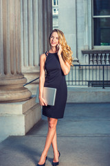 Obraz premium American Businesswoman working in New York. Wearing sleeveless black dress, high heels, holding laptop computer, a graduate student standing in vintage office building on campus, talking on phone..
