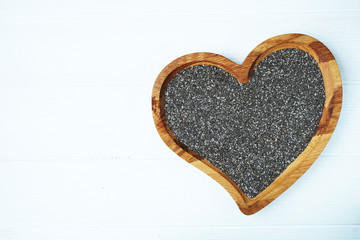 Wooden brown heart bowl full of chia seeds on white wooden background. Text space. Top view.
