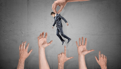 A giant hand holds a small businessman over many giant grabbing hands on concrete background.