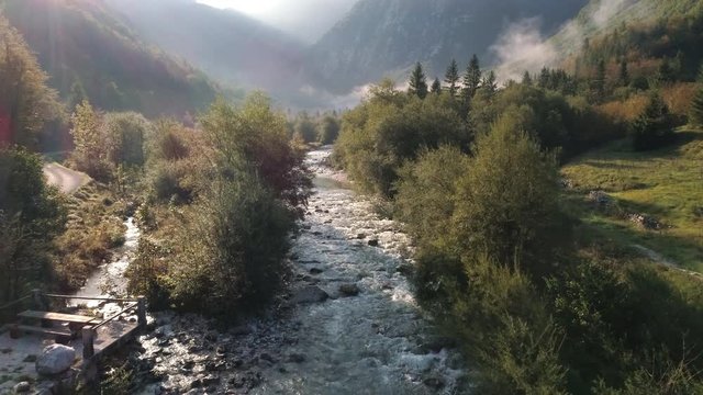 Flying past two people on small bridge over the Soca River in Lepena Valley as the Sun lights up the light foggy landscape through the canyon.
