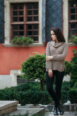 Beautiful young woman dressed in gray sweater and black pants posing in the city on a sunny day. Old town green garden and medieval windows background. Copy space