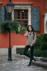 Beautiful young woman dressed in gray sweater and black pants posing in the old european city on a sunny day. Green garden and lamp post background.